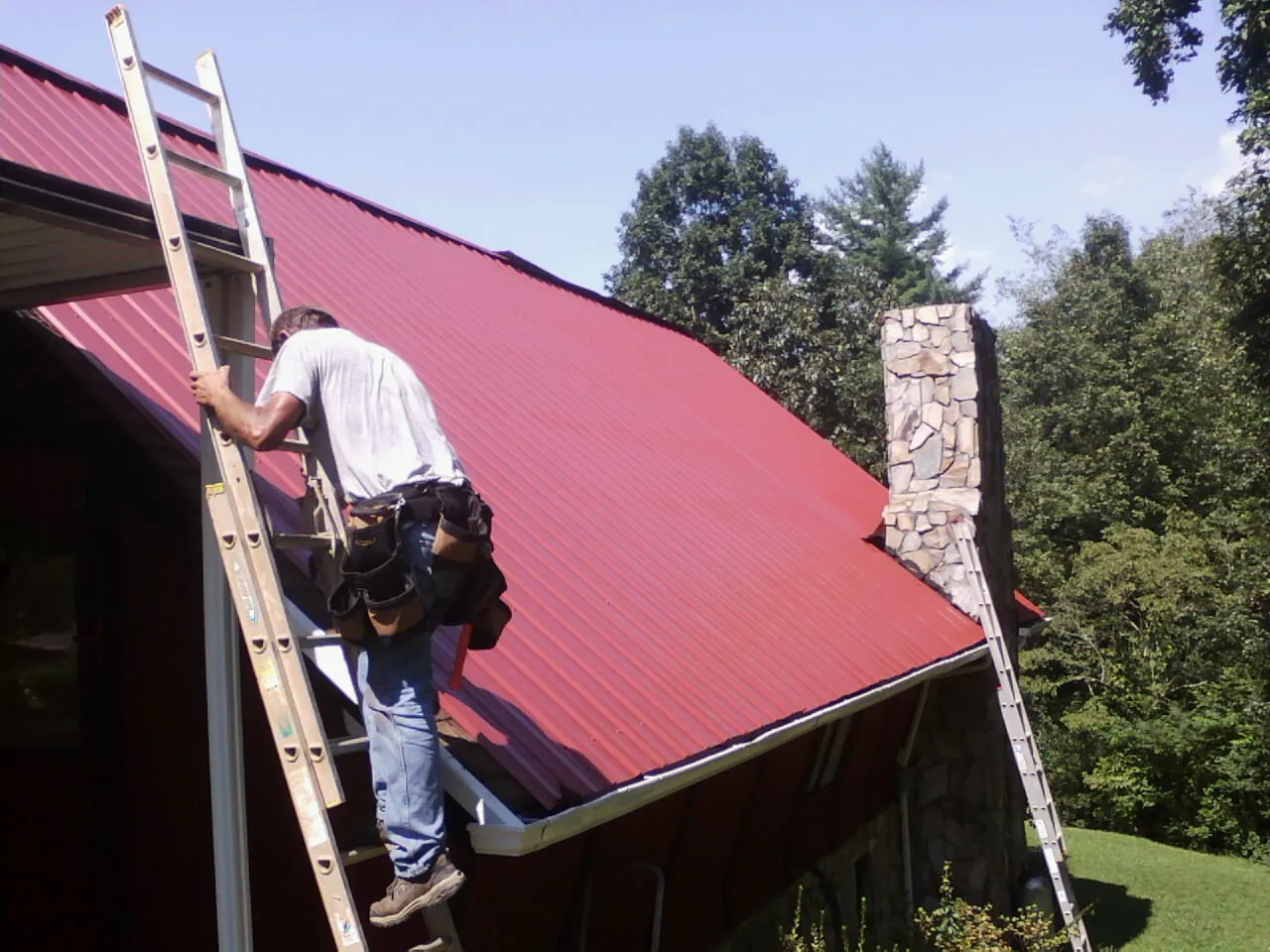 Roofer on a ladder beside a red metal roof and stone chimney — Soffit & Fascia Repair by Roofing Brothers in Twin Lakes